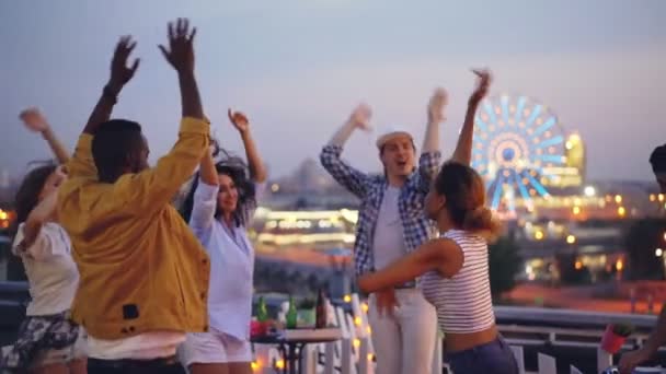 Zeitlupe fröhlicher Jugendlicher, die auf einer Open-Air-Party die Hände heben und sich entspannen, während ein DJ Musik mischt. schöne Stadt mit Abendlicht ist im Hintergrund zu sehen. — Stockvideo