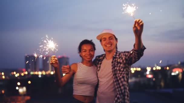 Retrato en cámara lenta de feliz pareja multirracial mujer afroamericana y hombre caucásico sosteniendo luces de bengala, mirando a la cámara y sonriendo de pie en la azotea . — Vídeos de Stock