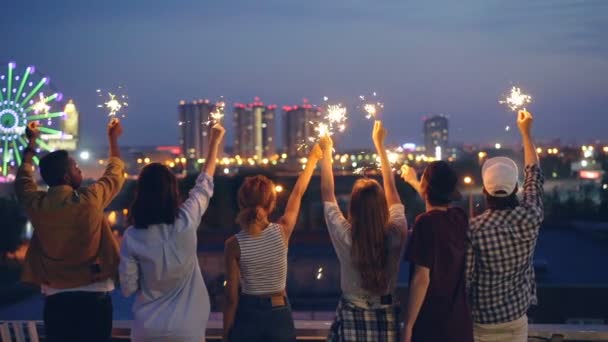 慢动作后视图的男性和女性持有孟加拉灯和移动举手站在屋顶上庆祝节日。友谊, 乐趣和夜生活概念. — 图库视频影像