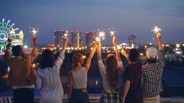 Visão traseira em câmera lenta de homens e mulheres segurando luzes de bengala e movendo as mãos levantadas em pé no telhado celebrando férias. Conceito de amizade, diversão e vida noturna . — Vídeo de Stock