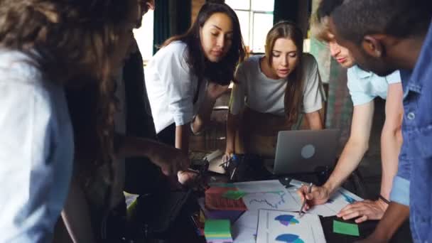 Groep jongeren collega's bespreekt grafieken en grafieken staande rond tafel in moderne kantoor. Business, teamwork en professionals concept. — Stockvideo