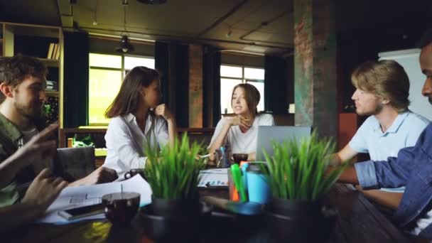 Une jeune femme s'entretient avec des collègues assis à table au bureau pour discuter de plans de travail pendant que son équipe l'écoute, un collègue masculin boit du thé . — Video