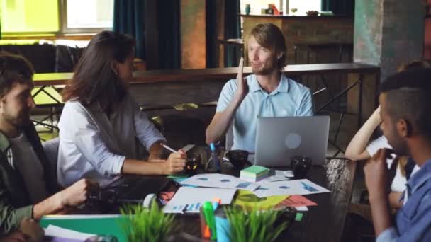 Cara alegre está contando uma história engraçada para seus colegas de trabalho e rindo sentado na mesa durante a pausa no local de trabalho. Juventude de sucesso, reunião de negócios e conceito de comunicação . — Vídeo de Stock