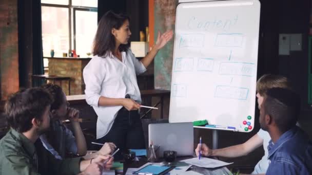 Jonge vrouw presentatie maakt voor haar collega's multiraciale groep, ze is permanent in de buurt van whiteboard, praten en wijzend op tekst aan boord. Bedrijfsleven en het onderwijs concept. — Stockvideo