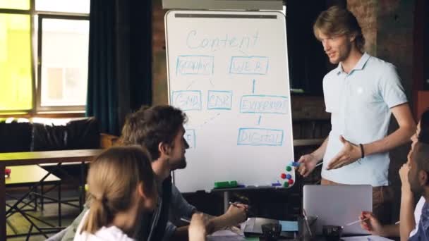 Pohledný mladý muž dělá kreativní prezentaci týmu pomocí tabule, ukazuje na text značky a mluví. Obchodní trenér a kancelářské koncept. — Stock video
