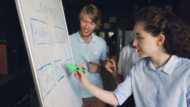 Jovens funcionários pessoas atraentes estão trabalhando com quadro branco olhando para gráficos, escrevendo com marcadores e falando de pé no escritório. Conceito de empresa, juventude e trabalho . — Vídeo de Stock