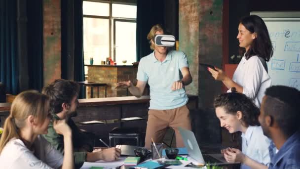 Young man wearing virtual reality glasses is having fun playing game while his colleagues are looking at him, laughing and using laptop and tablet in office. — Stock Video