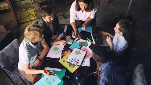 High angle view of multiracial team working with graphs and charts sitting at table, looking at papers and discussing strategy. Laptop, to-go coffee and documents are visible. — Stock Video