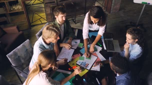 Groupe de travail multiracial analyse le diagramme et l'écriture sur des autocollants assis au bureau dans la salle de bureau. Vue en grand angle des gens qui parlent, pointent vers les graphiques et prennent des notes . — Video