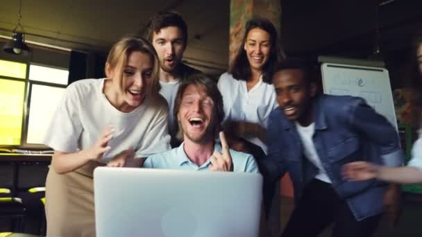 Захоплений молодий чоловік дивиться на екран ноутбука, радіє і виражає щастя, його колеги вітають його з успіхом, чіпляючись за руки і торкаючись плеча . — стокове відео