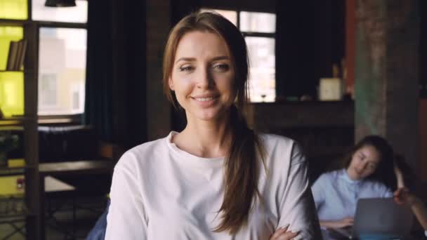 Portrait d'une jolie jeune employée de bureau debout sur le lieu de travail, regardant la caméra et souriant pendant que ses collègues travaillent en arrière-plan . — Video