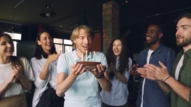 Ragazzo barbuto con viso espressivo sta esprimendo desideri e soffiando candele sulla torta di compleanno mentre i suoi colleghi applaudono e si congratulano con lui . — Video Stock