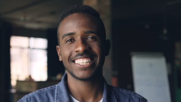 Primer plano retrato en cámara lenta del hombre afroamericano guapo de pie en la oficina de estilo loft, mirando a la cámara y sonriendo. Concepto de personas, trabajo y felicidad . — Vídeos de Stock