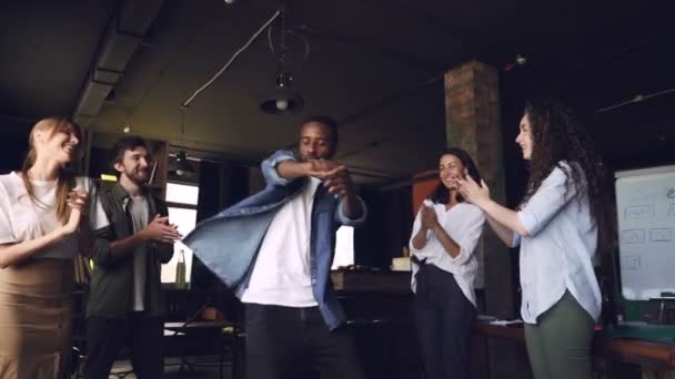 Медленное движение счастливого офисного работника афроамериканского парня, танцующего на корпоративной вечеринке, пока члены его команды хлопают в ладоши, смотрят на него и смеются . — стоковое видео