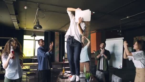 Movimento de sopro de jovem alegre dançando na mesa na festa do escritório com pilha de papel e jogando documentos enquanto sua equipe está rindo e dançando em torno dela . — Vídeo de Stock