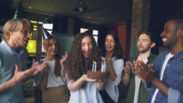 Cámara lenta de hermosa chica en partido sombrero celebración de la torta y soplar velas mientras sus compañeros de trabajo están de pie alrededor, aplaudiendo las manos y felicitándola en la oficina . — Vídeo de stock