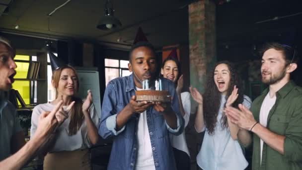Movimento lento de homem afro-americano animado soprando velas no bolo de aniversário e rindo enquanto sua equipe está parabenizando-o e batendo palmas no escritório . — Vídeo de Stock