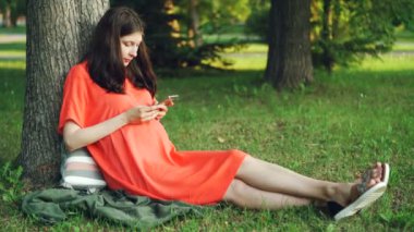 Casual giyim çekici hamile kadın parkta ağaç altında çimlerin üzerine oturan smartphone kullanıyor. Gebelik, insanlar, modern technogy ve gevşeme kavramı.