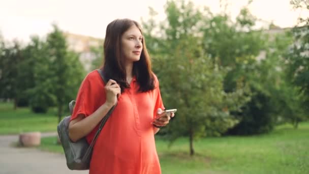 Menina grávida bonita esperando mãe está andando no parque da cidade e usando smartphone, jovem está segurando dispositivo e tela tocante, a natureza do verão é visível . — Vídeo de Stock