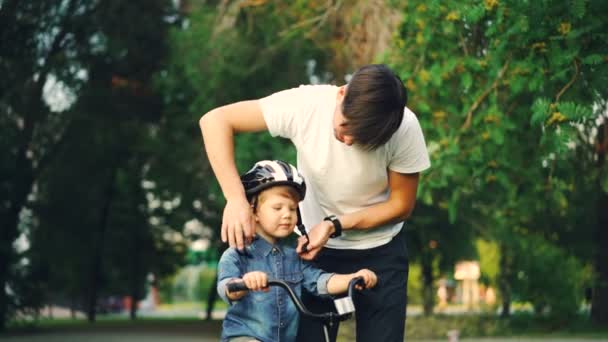 Pai carinhoso está colocando capacete de segurança em sua cabeça filhos pequenos, em seguida, ensinar menino feliz para andar de bicicleta, enquanto a mãe amorosa está assistindo-os e sorrindo . — Vídeo de Stock