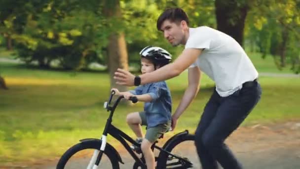 Schöner junger Mann liebevoller Vater bringt seinem kleinen Sohn bei, an Sommertagen im Park Fahrrad zu fahren, Junge fährt Fahrrad, während Papa ihn hält und rennt. — Stockvideo