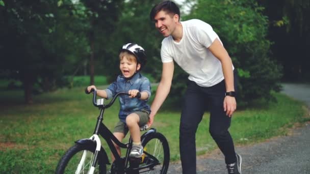 Slow motion of laughing child cycling in park with careful father who is teaching him to ride bicycle. Happy young family, fatherhood and childhood, active lifestyle concept. — Stock Video
