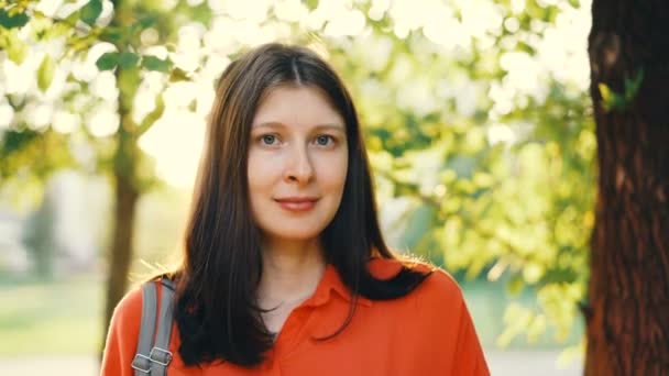 Close-up portrait of beautiful girl with brown hair looking at camera and smiling with summer park in background. Summertime, sunshine and relaxed people concept. — Stock Video