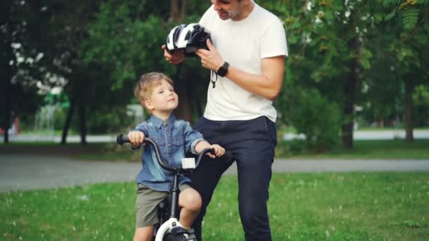Cámara lenta de papá cariñoso hablando con su hijo pequeño y luego poniendo casco de bicicleta protectora en la cabeza mientras el niño está sentado en bicicleta en el parque y sonriendo . — Vídeo de stock
