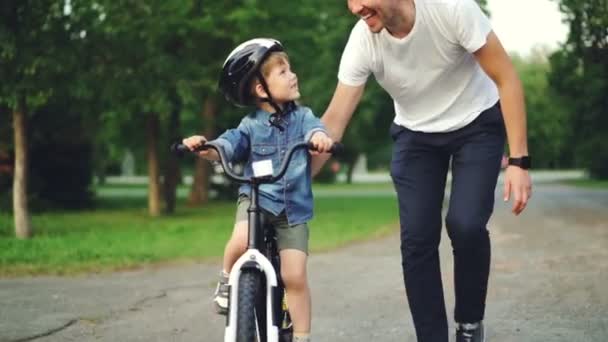 愛らしい息子公園自転車を押しながら子供に話で自転車に乗ることを教える愛情のあるお父さんのスローモーション。父権、幼年期およびアクティブなライフ スタイル コンセプト. — ストック動画