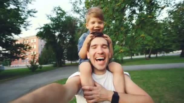 Boldog Apák szállító mosolygó fia, a nyak és videóinak selfie a smartphone kamera park séta során