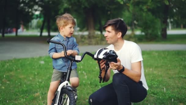 Starostlivý otec drží cyklistická přilba a mluvil s jeho rozkošný syn vysvětluje bezpečnostní předpisy, zatímco chlapec sedí na kole, s úsměvem a poslouchá tátu.