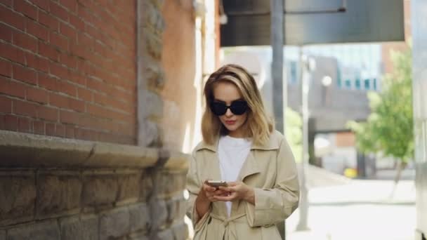 Mujer joven atractiva con el pelo rubio con abrigo de moda y gafas de sol está utilizando el teléfono inteligente caminando en la ciudad moderna. Gente, tecnología moderna y concepto juvenil . — Vídeo de stock