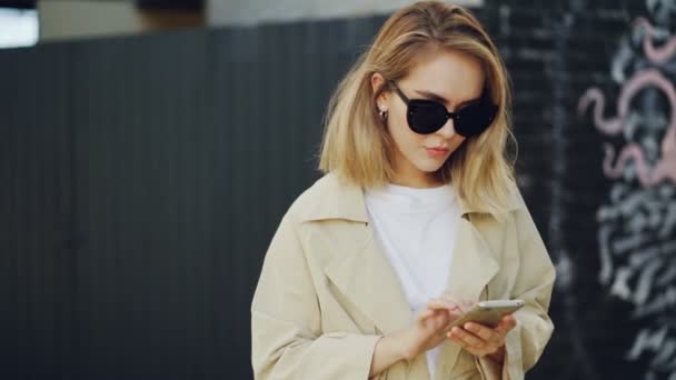 Красива блондинка використовує сенсорний екран смартфона, що стоїть на відкритому повітрі з графіті на стіні на фоні. Молода жінка носить сонцезахисні окуляри та літнє пальто . — стокове відео