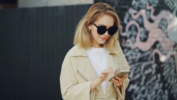 Smiling young lady is using smartphone browsing or chatting onine standing outdoors in modern city with graffiti in background. Girl is wearing sunglasses and coat. — Stock Video