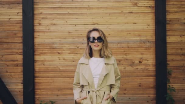 Retrato de una hermosa modelo de mujer joven con el pelo rubio de pie al aire libre, posando y mirando a la cámara con abrigo y gafas de sol. Concepto de personas, moda y belleza . — Vídeo de stock