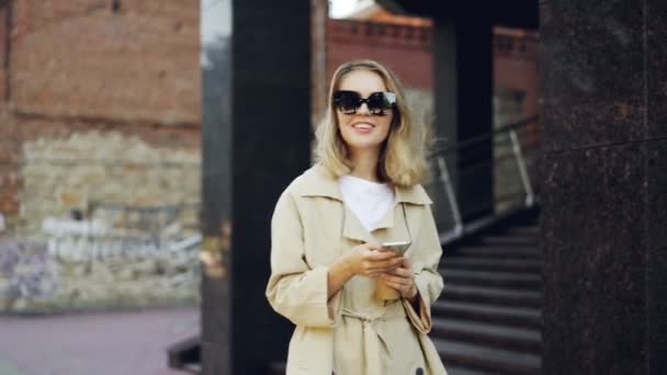 Elegant ung dam går längs gatan, med hjälp av smartphone och dricka till-go kaffe njuter promenad i vacker modern stad. Människor, modern livsstil och takeaway drycker koncept. — Stockvideo