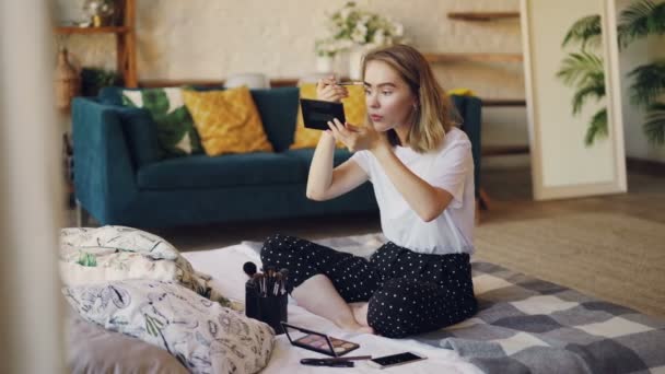 Hermosa chica rubia se está poniendo maquillaje pintando cejas con cepillo y kit de cosméticos sentado en la cama en casa. Belleza, mujeres jóvenes y el concepto de interiores modernos . — Vídeo de stock