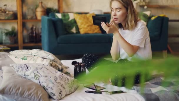 Linda loira é pintar lábios colocando maquiagem usando cosméticos decorativos profissionais sentados na cama em casa. Conceito de beleza, interiores e mulheres jovens . — Vídeo de Stock