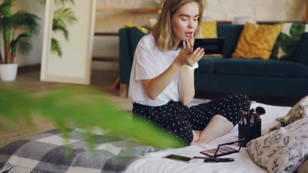 Jovencita atractiva se está poniendo maquillaje aplicando brillo de labios y luego mirándose en el espejo y sonriendo. Chica está sentado en la cama en casa con ropa casual . — Vídeos de Stock