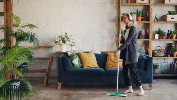 Beautiful young lady is listening to music through wireless headphones and doing housework cleaning the floor with mop. Girl is dancing and singing enjoying cleanup. — Stock Video