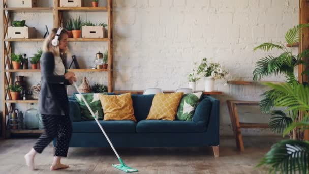 Pretty young woman is dancing and singing listening to music through headphones during housework at home, girl is dancing with mop moving around and having fun. — Stock Video