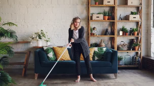 Mooi jong meisje huisvrouw is het schoonmaken van het huis en genieten van muziek, she's wearing koptelefoon, dansen en zingen thuis bedrijf mop tijdens opschonen. — Stockvideo