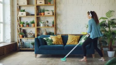 Neşeli genç kadın paspas ile sırasında rutin temizlik güzel loft tarzı daire içinde dans ediyor, şarkı ve kablosuz kulaklık ile müzik dinliyor.
