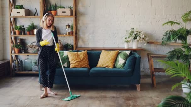 Attraente ragazza in cuffia è ascoltare musica e ballare wih mop durante il lavoro domestico, lei sta pulendo pavimento a casa e divertirsi. Concetto di donne, gioia e case . — Video Stock