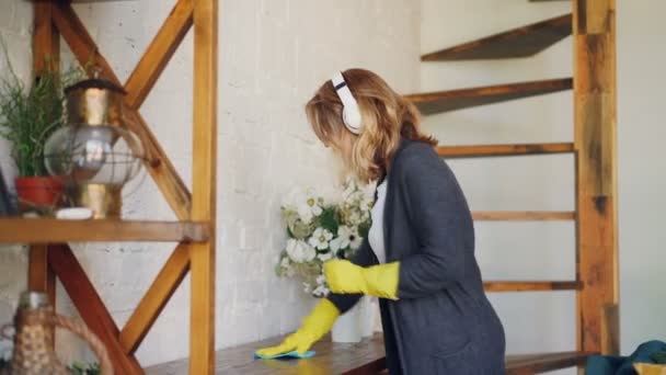 Mujer alegre está escuchando música con auriculares, bailando y cantando durante las tareas domésticas diarias. Chica está espolvoreando mesa sosteniendo tela y usando guantes . — Vídeos de Stock