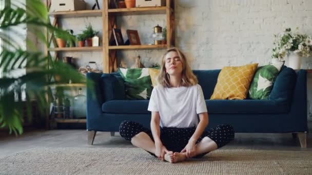 Menina bonita está relaxando em casa fazendo exercício de ioga sentado no chão com as mãos nos joelhos fechando os olhos e respirando dentro e fora. Meditação, relaxamento e conceito de pessoas . — Vídeo de Stock