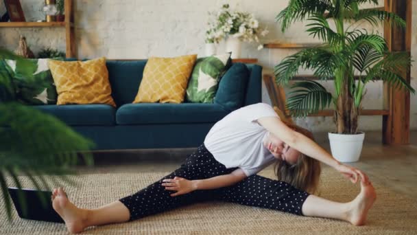 Elastyczne blondynka robi ćwiczenia rozciągające w domu siedzi na podłodze w nowoczesny apartament. Dziewczyna jest zginanie głowy do kolan, trzymając palce i sięga i nogi. — Wideo stockowe