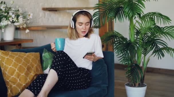 Menina bonita está usando smartphone, ouvir música através de fones de ouvido sem fio e beber chá sentado no sofá em casa. Tecnologia moderna e conceito millennials . — Vídeo de Stock