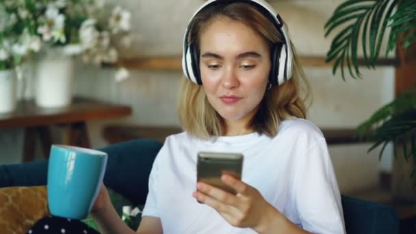 Счастливая барышня пользуется смартфоном и слушает музыку в беспроводных наушниках, сидя на диване в современной квартире. Технология и радость . — стоковое видео