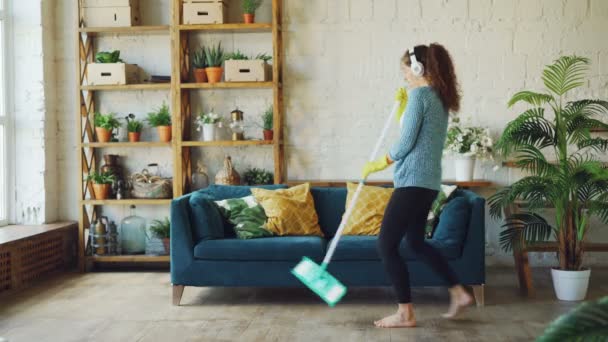 Grappige jonge dame is schoonmaken van het huis en luisteren naar muziek hoofdtelefoon dansen en zingen met platte mop dragen. Mooie meubels en groene planten zijn zichtbaar. — Stockvideo